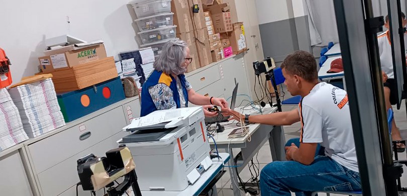 Fotografia de uma mulher, que veste o colete amarelo e azul da Justiça Eleitoral, coletando as d...