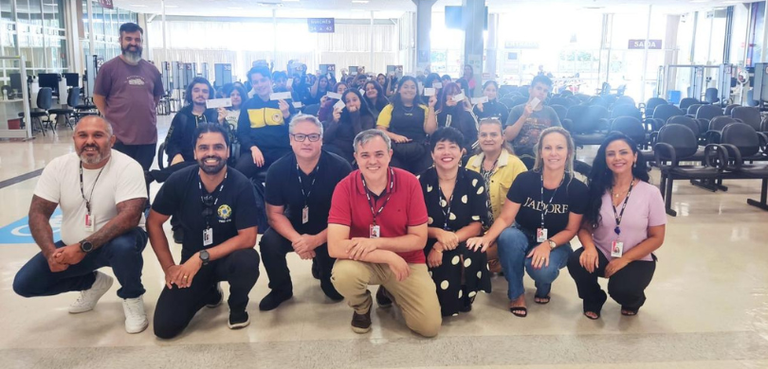 Fotografia de um grupo de pessoas em um ambiente institucional amplo. Em primeiro plano, oito ad...
