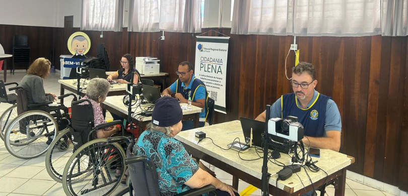 14ª ZE de Ponta Grossa realiza cadastramento de eleitores idosos em instituições de longa permanência Fotografia que retrata um ambiente interno de atendimento ao público, onde três homens vestindo ...