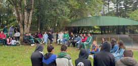 Na imagem, há várias pessoas sentadas em um local ao ar livre, como se fosse um meio círculo. No...