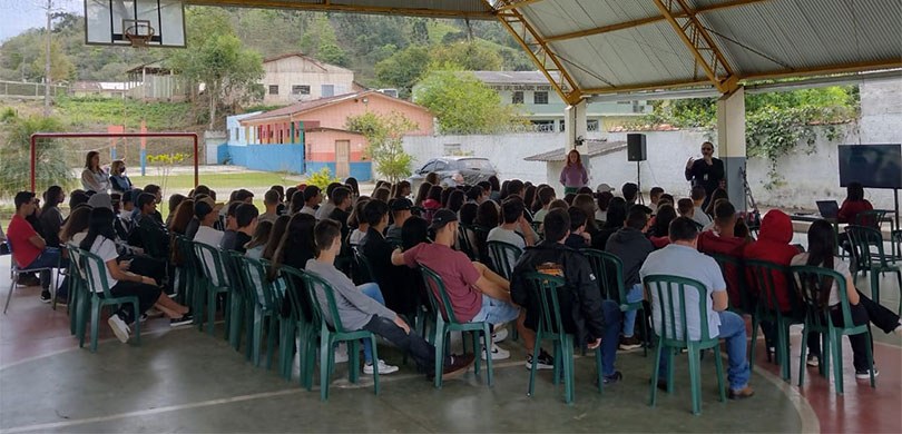 Fotografia de uma quadra de futebol de uma escola. Estudantes aparecem sentados em cadeiras de p...