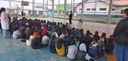 Fotografia de dezenas  de crianças uniformizadas sentadas no chão da quadra de uma escola, elas ...