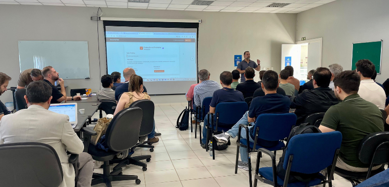 Fotografia de um homem falando diante de uma plateia, formada majoritariamente por homens, senta...
