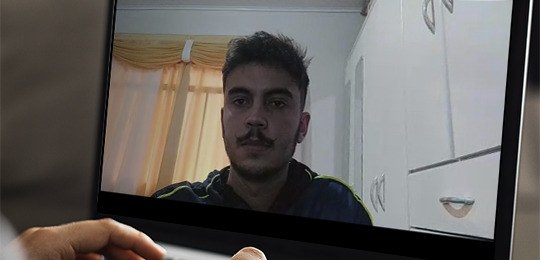 Foto do acadêmico João Gabriel Cordeiro Cruz sendo exibida em uma tela de notebok e uma mão sobr...