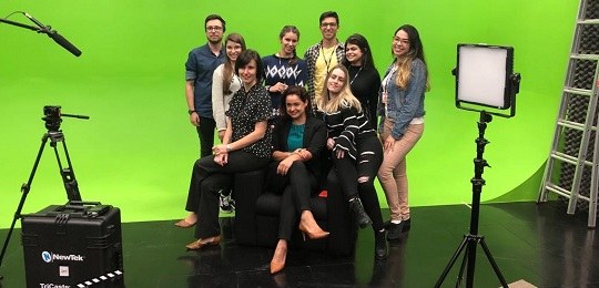 Na imagem nove pessoas posando para foto à frente de uma parede verde em um estúdio de TV. Seis ...