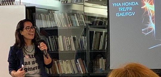 2019-10-24-Yna Mulher segurando um microfone, ao fundo livros.