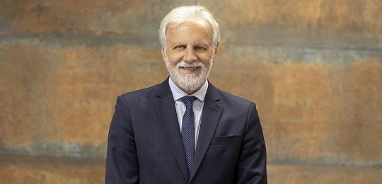 Fotografia da cintura para cima um senhor de terno azul-marinho e gravata sobre um fundo de pare...