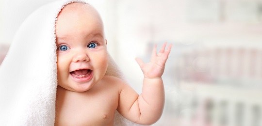 Fotografia de um bebê de olhos azuis sem roupa com uma tolha branca na cabeça. Ele sorri e levan...