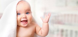 Fotografia de um bebê de olhos azuis sem roupa com uma tolha branca na cabeça. Ele sorri e levan...