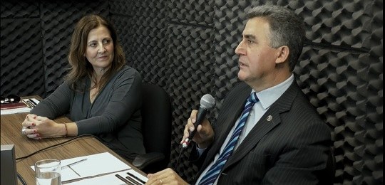 Fotografia de duas pessoas dentro de um estúdio. À esquerda, há uma mulher com os braços sobre a...