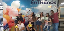 pessoas com placas sorrindo em frente a um painel com baloes escrito entrenós