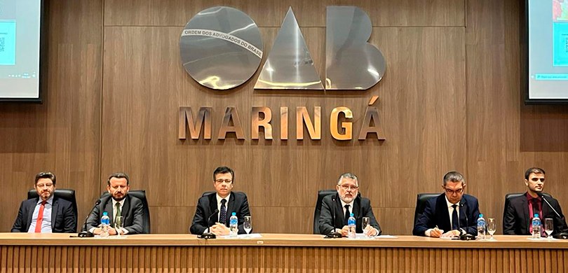 Fotografia de seis homens de terno e gravata sentados atrás de uma bancada com microfones. Ao fu...
