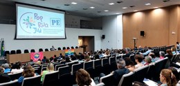Fotografia de um auditório com platéia presente. No palco, há um homem atrás de uma bancada. Aci...