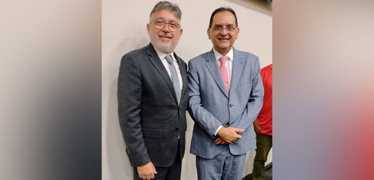 Fotografia de dois homens posando sorrindo de pé. Eles vestem terno e gravata e usam óculos. Ao ...