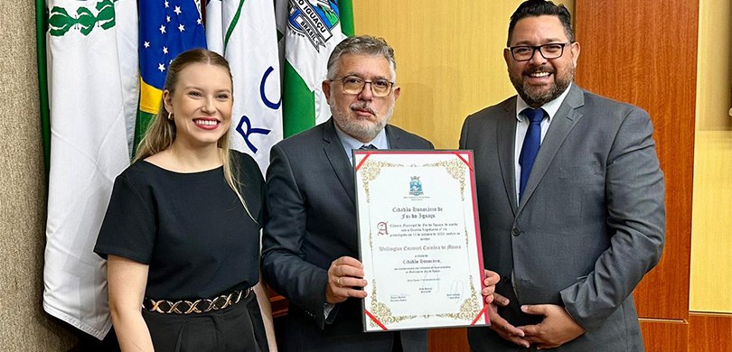 Fotografia de uma mulher de vestido preto e dois homens de terno. O homem ao centro segura um ce...