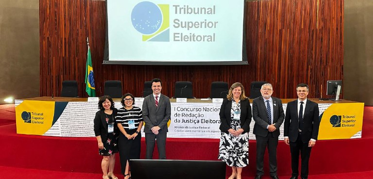 2023-11-23-concurso Fotografia de três mulheres e três homens posando sorrindo em frente a um telão em que se lê: Tr...