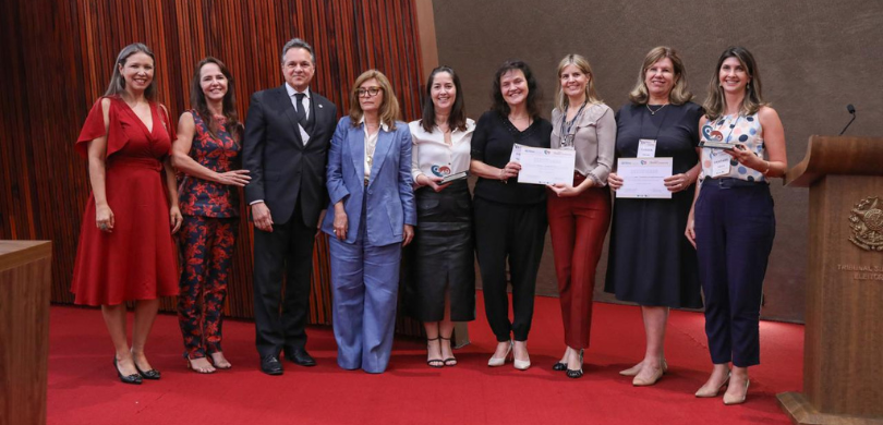 Fotografia de um homem de terno e oito mulheres, que vestem roupas formais, posando sorrindo no ...