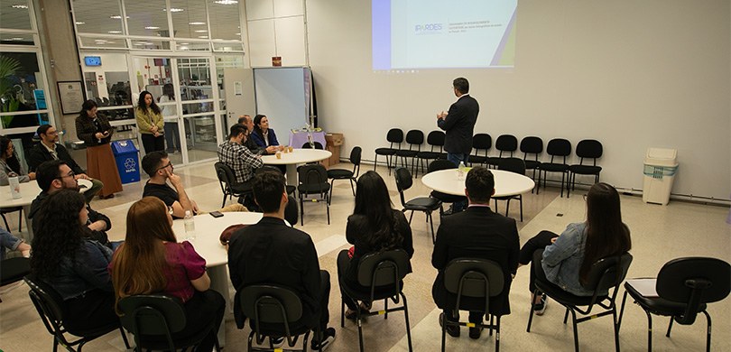 Fotografia em que são vistas várias pessoas sentadas de contas observando um homem em pé falando...