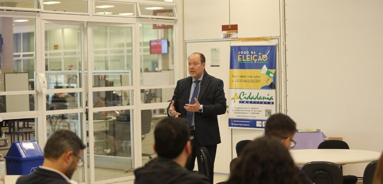 Fotografia em um homem vestindo terno escuro aparece em pé falando ao público. Em primeiro plano...