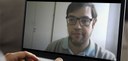Fotografia da tela de um notebook, durante videoconferência. Um homem de cabelos escuros e que u...