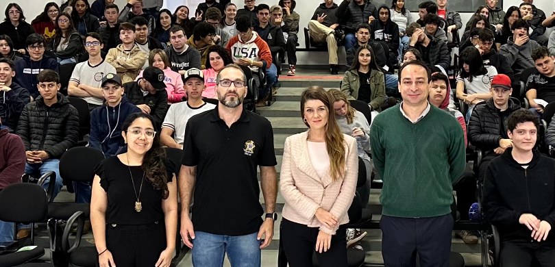 Em um auditório, há dois homens e duas mulheres de pé, olhando para a câmera. Atrás deles vários...