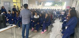 Fotografia de um homem de camisa social cinza e calça jeans, que aparece de costas, palestrando ...