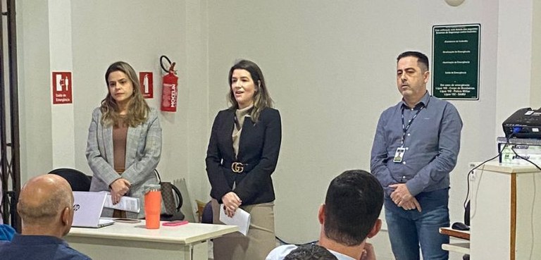 Fotografia de duas mulheres e um homem em pé durante uma palestra. A sala tem paredes brancas co...