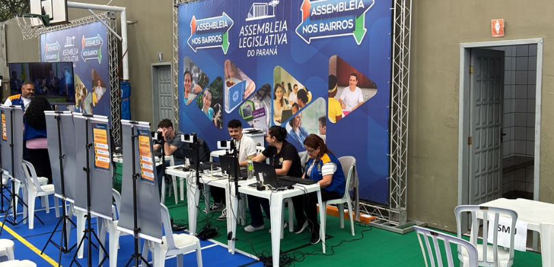 Fotografia dos atendentes da Justiça Eleitoral sentados atrás de guichês de atendimento. Ao fund...