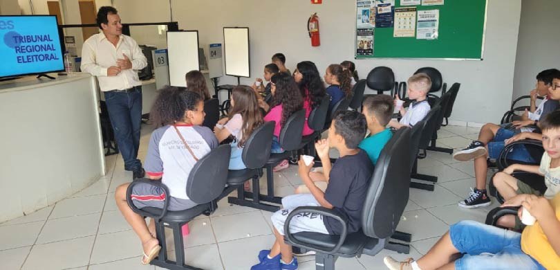 Fotografia de um homem em pé falando diante de crianças, que estão sentadas em cadeiras pretas, ...