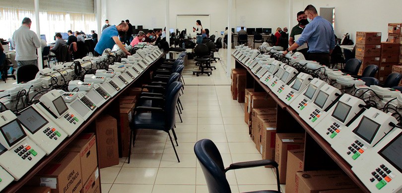 A menos de cem dias das eleições, Justiça Eleitoral testa sistemas da urna em Curitiba Fotografia de duas mesas com urnas eletrônicas enfileiradas, uma ao lado da outra. Algumas pesso...