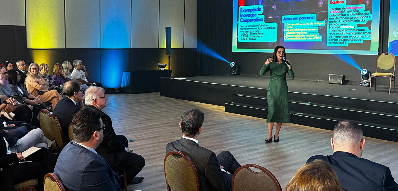 Fotografia de uma mulher de vestido verde, que segura um microfone com a mão esquerda, enquanto ...