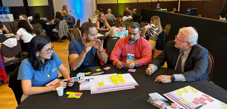 Fotografia de três homens e uma mulher sentados em cadeiras marrons ao redor de uma mesa preta, ...
