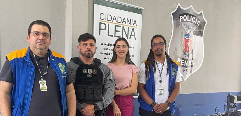 Fotografia em que aparecem três homens, dois vestindo coletes da Justiça Eleitoral e um policial...