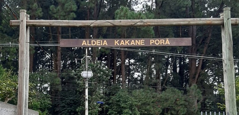 Fotografia de uma placa de madeira escrito em amarelo: Aldeia Kakané Porã. Ao fundo, diversas ár...