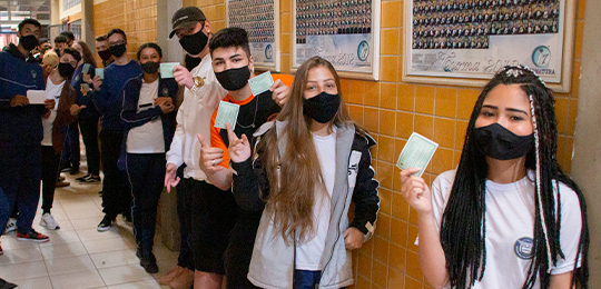 No corredor de uma escola, um grupo de jovens exibe seu primeiro título, impresso, para a câmera...
