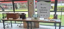 Fotografia em que se observa uma mesa retangular de madeira com diversos itens organizados sobre...