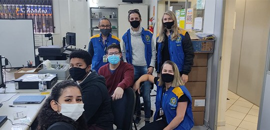 Foto de sete pessoas. Todas estão de máscara. Três estão em pé, três estão sentadas em cadeiras ...