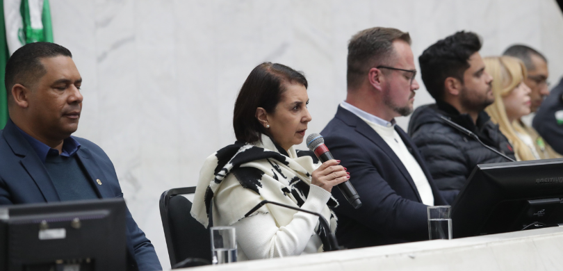 Fotografia de uma mesa de autoridades, composta por homens e mulheres. Em destaque, uma mulher d...