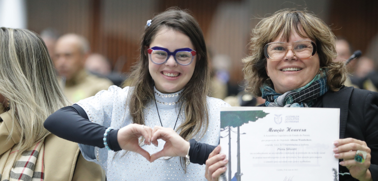 Fotografia de uma jovem, que faz um coração com as mãos, posando sorrindo ao lado de uma mulher,...