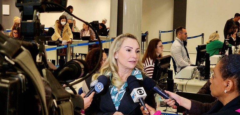 Fotografia de uma mulher de cabelos loiros cercada por microfones e repórteres. À direita, em pr...