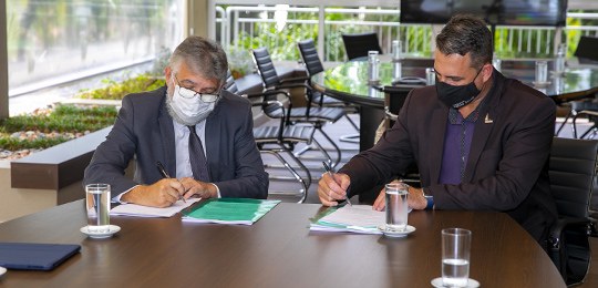 Fotografia de dois homens sentados ao redor de uma mesa assinando um documento cada. Ao fundo, h...