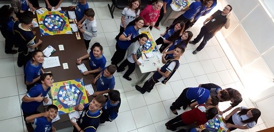 Fotografia tirada de cima de vários grupos de estudantes posicionados ao redor de mesas. Sobre a...