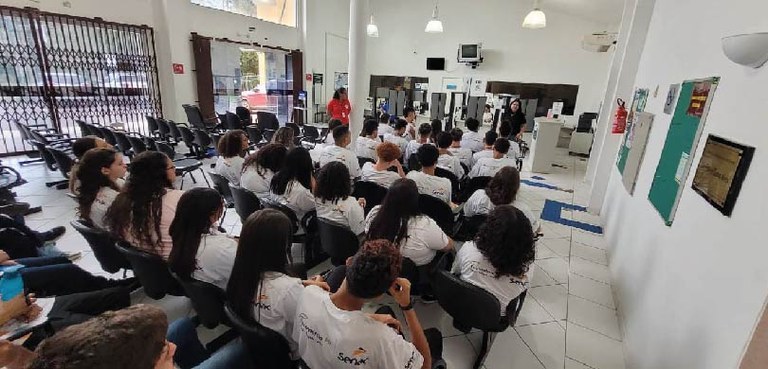 Foto tirada do fundo de uma sala com paredes brancas, em que estudantes vestidos com o uniforme ...