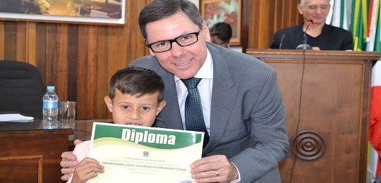 Fotografia de um homem de terno abaixado ao lado de uma criança. Eles seguram um diploma juntos.