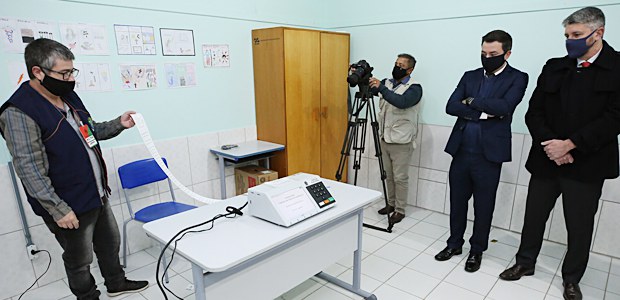 Foto de três homens de máscara olhando para uma urna eletrônica imprimindo a zerésima e um homem...