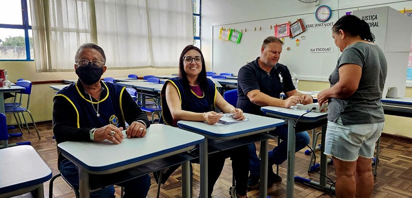 Três pessoas estão sentadas em carteiras escolares. Ao fundo uma sala de aula com outras carteir...