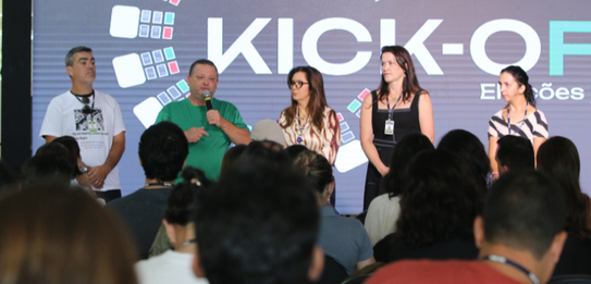 Fotografia de nove pessoas, entre homens e mulheres, posando sorrindo em frente a um edifício co...