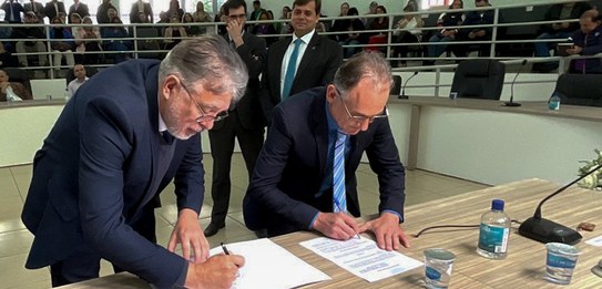 Fotografia de quatro homens sentados atrás de uma mesa de autoridades. Todos usam terno. Ao redo...
