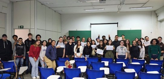 Fotografia de uma mulher em pé palestrando para jovens uniformizados em uma sala de aula com par...
