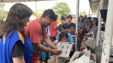 Duas pessoas adultas, uma mulher de colete azul e um homem
de camisa vermelha, interagem com um...